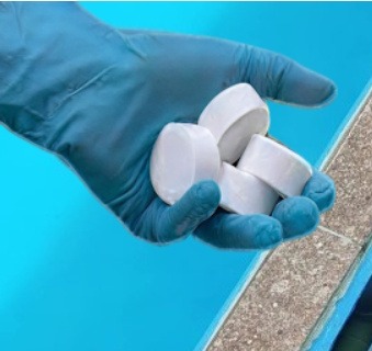 Gloved hand holding silver nanoparticle AgNP pool disinfection tablets over blue pool water.