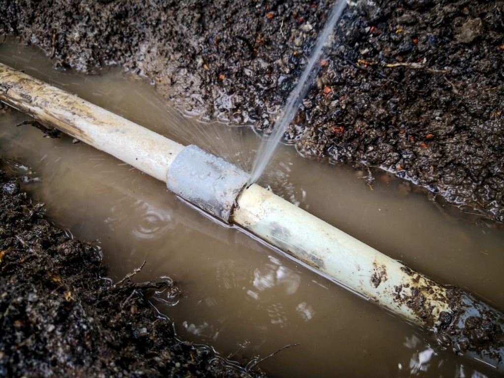 A close-up of a broken white underground pool pipe aggressively spraying water into a muddy dirt trench.