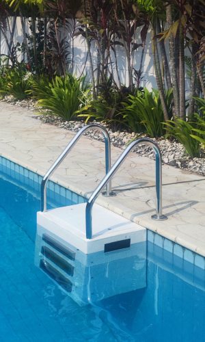 Side view of a white plug and play pool filter box mounted to the wall of a tiled swimming pool with stainless steel handrails.