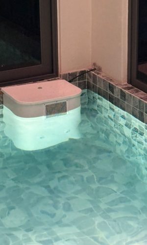 Illuminated white plug and play pool filter box mounted in a tiled indoor pool corner at night.