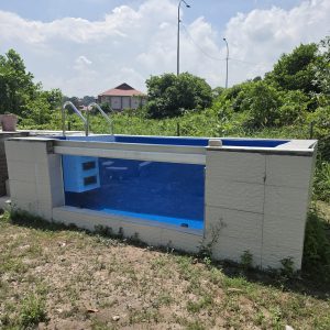 Exterior front-facing view of a custom glass-window swimming pool featuring an internal blue plug and play filtration system.