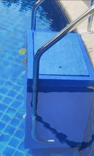 Side angle view of a blue plug and play pool filter casing with stainless steel handrails submerged along a tiled pool edge.