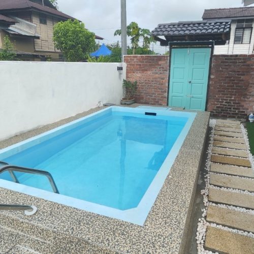 A light blue rectangular fiberglass pool with a raised pebble edge. It sits between a white boundary wall and a traditional wooden house, featuring stepping stones and white garden pebbles.
